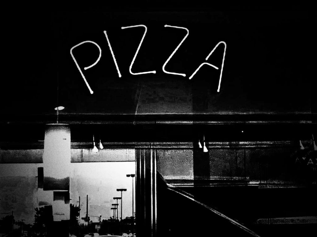 A black and white photo of a neon sign that reads "pizza" outside of a pizzeria.
