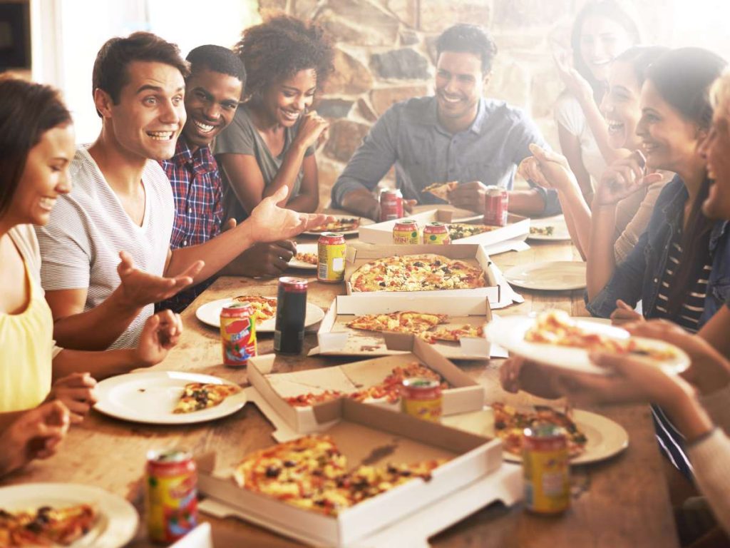 A group of people eating pizza and drinking around a table at a pizza party.